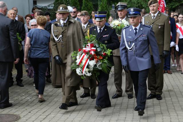 Przedstawiciele służb mundurowych składający wiązankę kwiatów pod tablicą upamiętniającą okrutne wydarzenia.