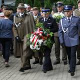 Przedstawiciele służb mundurowych składający wiązankę kwiatów pod tablicą upamiętniającą okrutne wydarzenia.