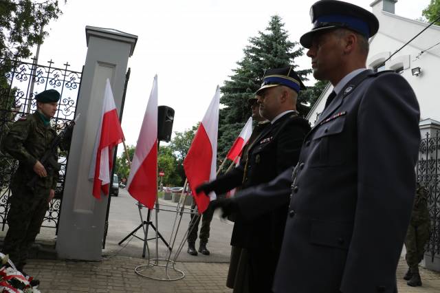 Przedstawiciele służb mundurowych składający wiązankę kwiatów pod tablicą upamiętniającą okrutne wydarzenia.