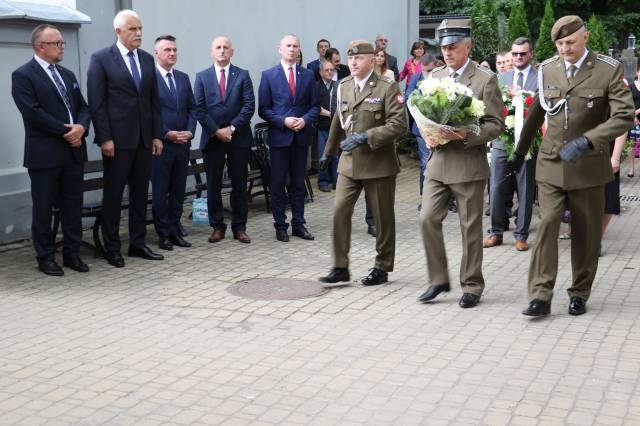 Przedstawiciele służb mundurowych składający wiązankę kwiatów pod tablicą upamiętniającą okrutne wydarzenia.